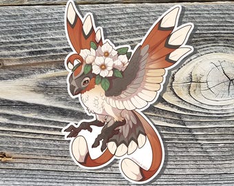 May include: A sticker featuring a stylized bird with outstretched wings, adorned with a floral crown. The bird has brown, tan, and white feathers, a gray face, and a long, curved tail. The sticker is on a wooden background.
