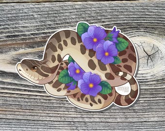 May include: A sticker featuring a brown and white snake with a curled tail, surrounded by purple flowers with green leaves. The sticker has the text "Imanika" and the year "2022" in the bottom right corner.