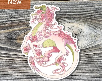 May include: A die-cut sticker featuring a mythical dragon creature with pale pink fur, a light green underbelly, and long flowing mane. The dragon is adorned with clusters of pink flowers and green leaves, depicted in a dynamic pose with a long tail.