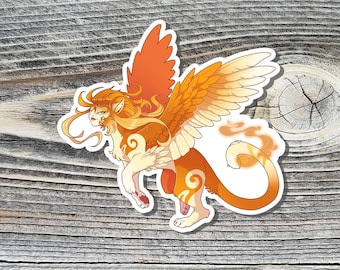 May include: A sticker featuring a mythical creature with orange and cream coloring. The creature has large feathered wings, a lion-like body, and a human-like face with long flowing hair. The sticker is on a wooden surface.