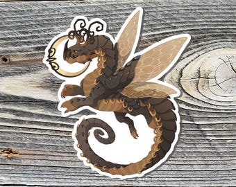 May include: A sticker featuring a stylized dragon with brown and tan scales, wings, and a crescent moon. The dragon has a long, curled tail and is set against a wooden background.