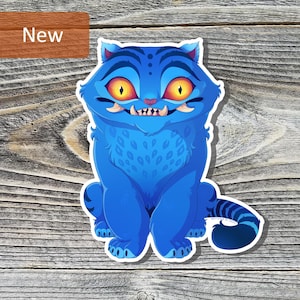 Puede incluir: Adhesivo de gato de dibujos animados azul con grandes ojos amarillos y dientes afilados. El adhesivo tiene un borde blanco y una cola a rayas. La palabra "New" está en un rectángulo marrón en la esquina superior izquierda.