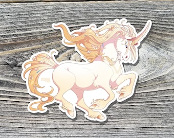 May include: A sticker featuring a cream-colored unicorn with a flowing mane and tail in shades of peach and gold. The unicorn has a long, spiraled horn and is decorated with small flowers. The sticker is on a wooden surface.