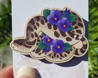 May include: A wooden sticker featuring a snake with a tan and brown spotted pattern, adorned with purple flowers and green leaves. The snake is coiled, and the sticker is on a white background.