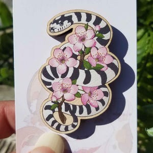 May include: A wooden cutout of a black and white striped snake with pink cherry blossoms. The snake is curled around the flowers, and the blossoms are in full bloom.