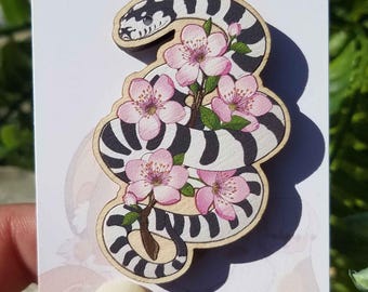 May include: A wooden cutout of a black and white striped snake with pink cherry blossoms. The snake is curled around the flowers, and the blossoms are in full bloom.