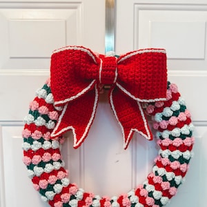 May include: A handmade crochet wreath with a large red bow. The wreath is a ring of alternating red, pink, green, and white crochet bobbles. The bow is red with white trim and is attached to the top of the wreath.