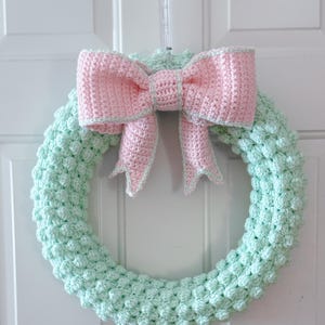 May include: A handmade crochet wreath with a light green base and a large pink bow. The wreath is round and has a textured, bobbled appearance. The bow is centered at the top and has a light green trim.