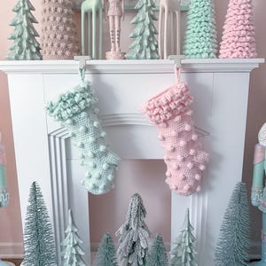 May include: Two crocheted Christmas stockings hanging on a white fireplace mantel. The stocking on the left is light blue with white pom-poms and the stocking on the right is pink with white pom-poms. There are several small artificial Christmas trees on the mantel and below the fireplace.