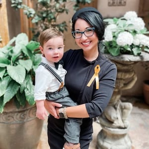 May include: A toddler wearing a white collared shirt, a black bow tie, grey trousers with suspenders, and brown shoes, held by an adult wearing a black top with a yellow awareness ribbon pinned to the chest and black-rimmed eyeglasses.