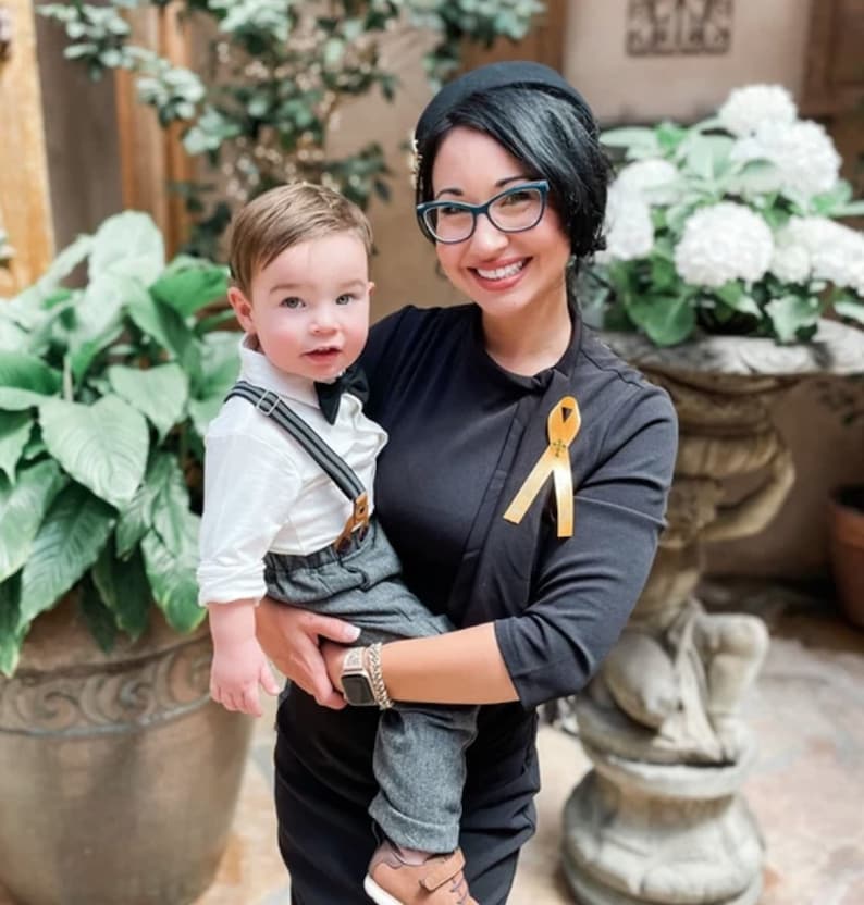 May include: A toddler wearing a white collared shirt, a black bow tie, grey trousers with suspenders, and brown shoes, held by an adult wearing a black top with a yellow awareness ribbon pinned to the chest and black-rimmed eyeglasses.