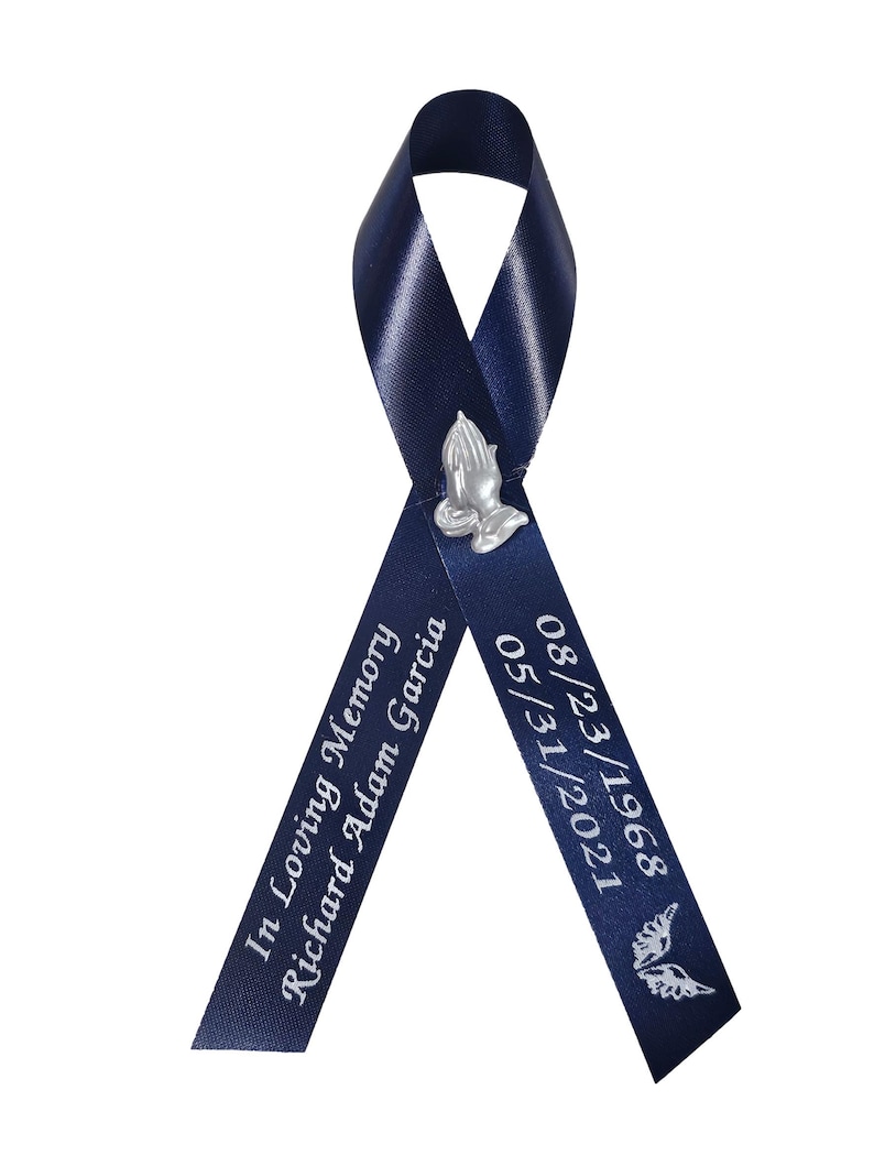 May include: A navy blue satin ribbon folded into a loop, featuring a silver-toned praying hands charm at the center. The left tail is printed with "In Loving Memory Richard Adam Garcia" and the right tail with "08/23/1968 05/31/2021" and a dove.