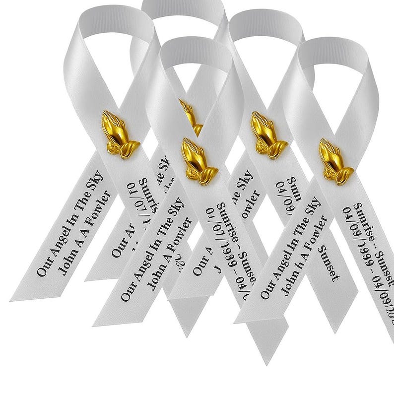 May include: A group of white awareness ribbons featuring a gold-toned praying hands emblem. Each ribbon is printed with the text Our Angel In The Sky, John A A Fowler, and the dates Sunrise 04/07/1999 - Sunset 04/09/2024 in black lettering.