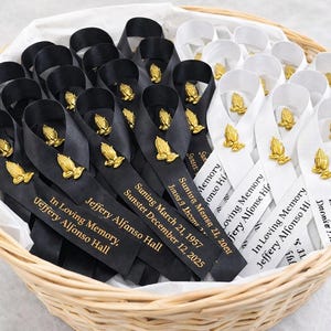 May include: A collection of black and white satin ribbons arranged in a woven basket. Each ribbon features a gold-toned praying hands emblem and is printed with the text In Loving Memory, Jeffery Alfonso Hall, and specific dates.