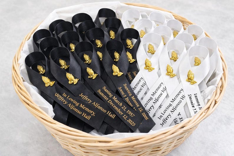 May include: A collection of black and white satin ribbons arranged in a woven basket. Each ribbon features a gold-toned praying hands emblem and is printed with the text In Loving Memory, Jeffery Alfonso Hall, and specific dates.