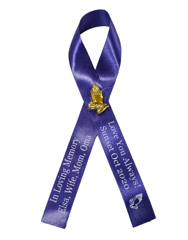 May include: A purple awareness ribbon featuring a gold-toned praying hands pin at the center. The left tail is printed with "In Loving Memory Elsa, Wife, Mom, Oma" and the right tail features "Love You Always! Sunset Oct 2020" with a small icon.