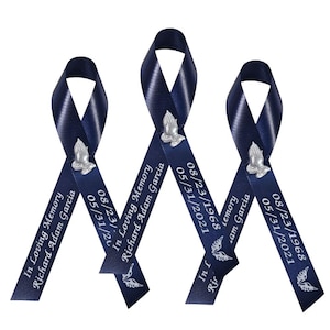 May include: Three dark blue satin ribbons arranged in a vertical loop design. Each ribbon features a silver-toned praying hands emblem, the text "In Loving Memory Richard Adam Garcia," the dates "08/23/1968" and "05/31/2021," and a small angel wing icon.