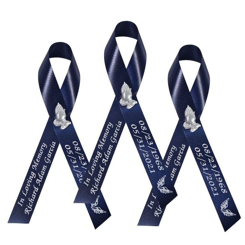 May include: Three dark blue satin ribbons arranged in a vertical loop design. Each ribbon features a silver-toned praying hands emblem, the text "In Loving Memory Richard Adam Garcia," the dates "08/23/1968" and "05/31/2021," and a small angel wing icon.