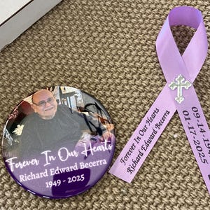 May include: A circular memorial button featuring a photograph of a person, the text Forever In Our Hearts, Richard Edward Becerra, and 1949-2025, paired with a lavender ribbon printed with the same text, dates, and a small silver cross charm.