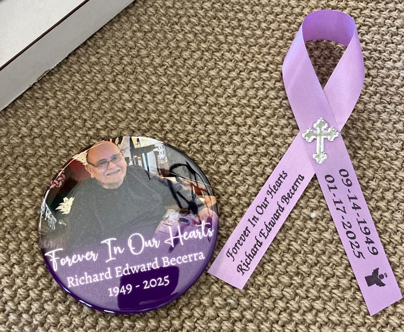 May include: A circular memorial button featuring a photograph of a person, the text Forever In Our Hearts, Richard Edward Becerra, and 1949-2025, paired with a lavender ribbon printed with the same text, dates, and a small silver cross charm.