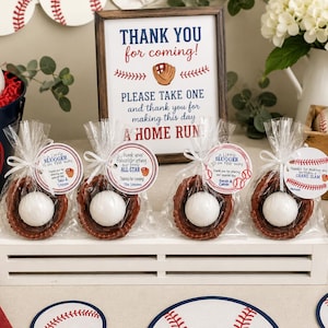 May include: Four clear plastic favour bags each containing a brown baseball glove holding a white baseball. Each bag is tied with a white ribbon and a round paper tag featuring baseball graphics and text including A Little Slugger is on the way.
