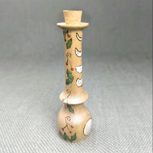 Peut inclure: Un bouchon de bouteille en bois avec un bouchon en liège et un motif peint de feuilles de houx et de lunes blanches. Le bouchon de bouteille est peint dans des tons de brun, vert, blanc et rouge.