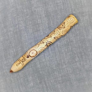 Puede incluir: Una varita de madera con una punta puntiaguda y un diseño tallado de varios símbolos, incluyendo un pentagrama, un ankh y un patrón en espiral.