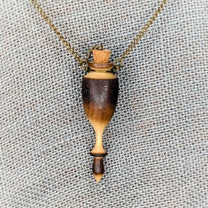 May include: A wooden pendant necklace with a brown and light wood finish. The pendant is shaped like a bottle with a cork stopper. The necklace is made of a brass chain.