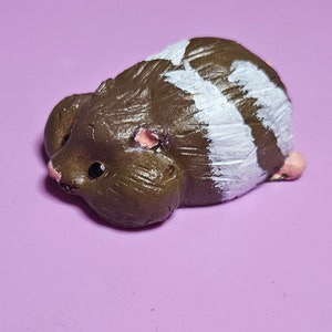 May include: A small brown and white hamster figurine with pink ears and a black eye. The figurine is lying on its side with its head turned to the left.