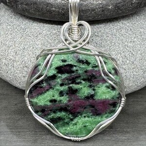 May include: A handcrafted pendant featuring a polished, oval-shaped stone with a vibrant mix of green, black, and maroon hues. The stone is encased in a delicate, silver-colored wire wrap, suspended from a matching chain. The pendant's design is intricate and eye-catching.