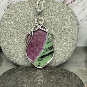 May include: A silver wire wrapped pendant featuring a green and pink gemstone. The pendant is on a silver chain and is resting on a gray stone.