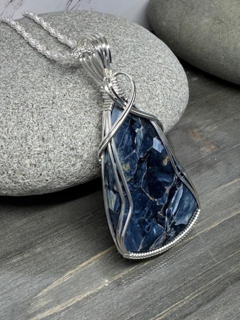 May include: A silver wire wrapped pendant featuring a blue and white tiger's eye gemstone. The pendant is hanging from a silver chain.
