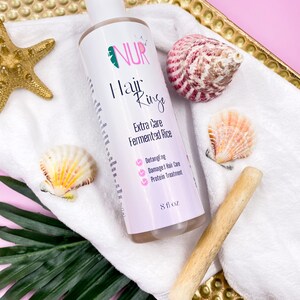 Op de afbeelding: Een witte fles NUR Hair Rinse Extra Care Fermented Rice, met een roze etiket en een groen blaadjeslogo. De fles staat op een witte handdoek met schelpen en een houten stok. Het etiket somt de voordelen van het product op: Ontwarren, Beschadigde Haarverzorging en Prote&iuml;nebehandeling. De fles bevat 8 fl oz.