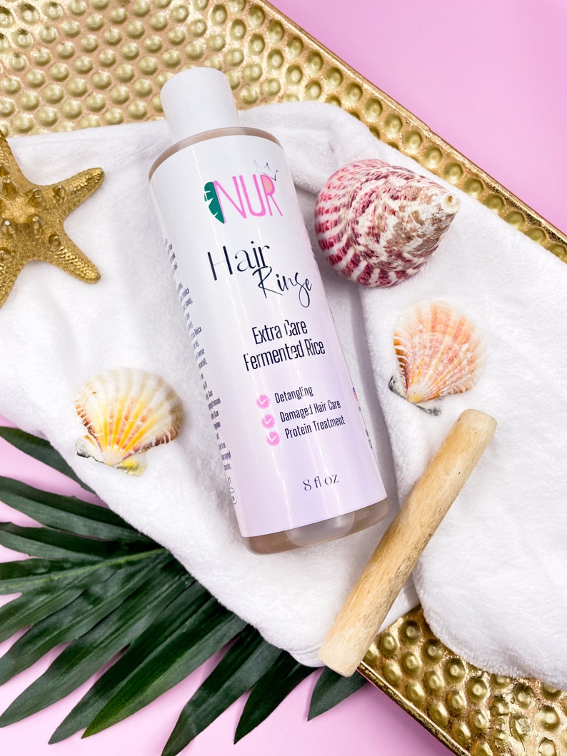 Op de afbeelding: Een witte fles NUR Hair Rinse Extra Care Fermented Rice, met een roze etiket en een groen blaadjeslogo. De fles staat op een witte handdoek met schelpen en een houten stok. Het etiket somt de voordelen van het product op: Ontwarren, Beschadigde Haarverzorging en Prote&iuml;nebehandeling. De fles bevat 8 fl oz.