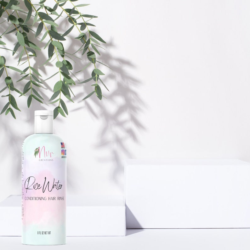 Op de afbeelding: Een witte fles met een roze etiket waarop "Rice Water Conditioning Hair Rinse" en de merknaam "Nuru Creations" staat. De fles staat op een wit oppervlak met een groene, bladerrijke tak op de achtergrond.