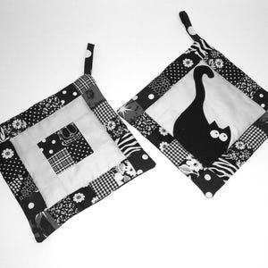 Puede incluir: Dos agarraderas acolchadas en blanco y negro. Una presenta una silueta de gato negro, la otra un diseño de patchwork con estampados florales, lunares y geométricos. Cada una tiene un lazo para colgar.