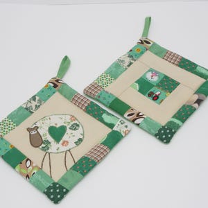 Può includere: Due presine trapuntate con disegni patchwork verdi e beige. Una presenta una pecora con un motivo floreale a forma di cuore, l'altra un motivo quadrato con un trattore e un orologio. Entrambe hanno anelli verdi per appenderle.