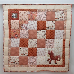 Op de afbeelding: Een patchworkquilt met een boerderijdierenthema. De quilt heeft vierkanten met verschillende patronen en kleuren, waaronder oranje, crème en bruin. Op de quilt is een bruin paard geappliqueerd. De quilt is omrand met een crèmekleurige stof.