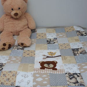 Puede incluir: Una colcha de retazos con un oso de peluche marrón, un pájaro y varios patrones en beige, marrón y gris. La colcha está hecha de cuadrados de diferentes telas, con patrones que incluyen nubes, flores y guijarros.