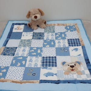 Puede incluir: Una colcha patchwork para bebé en tonos azules y blancos, con diversos patrones como ositos de peluche, peces y corazones. Un osito de peluche de peluche está encima de la colcha. La colcha tiene un borde azul claro y mide aproximadamente 100 x 100 cm.