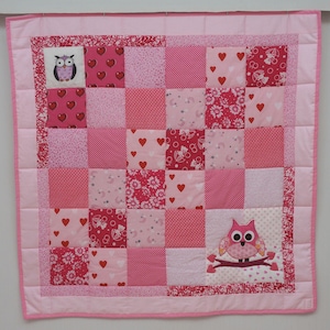 May include: A pink and white patchwork quilt with a variety of patterns, including hearts, flowers, and polka dots. Two squares feature owl appliqués. The quilt is bordered with a solid pink fabric and pink binding.