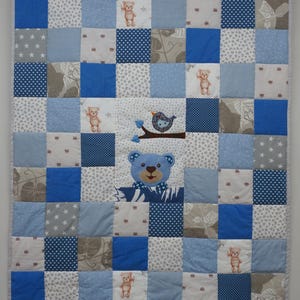 Puede incluir: Una colcha de retazos con una cuadrícula de cuadrados de tela azules, grises y blancos. El diseño incluye patrones de estrellas, corazones y ositos de peluche. Una aplicación central muestra la cara de un osito azul y un pájaro en una rama.