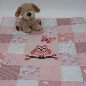 Op de afbeelding: Een patchwork deken in roze en witte tinten met een centrale applicatie van een uil op een tak met daarboven een vlinder. De deken is samengesteld uit diverse vierkanten met patronen zoals sterren, stippen en uilmotieven, inclusief een beige pluche hond.
