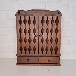 May include: A vintage wooden cabinet with two doors and two drawers. The doors feature a decorative pattern of oval cutouts. The cabinet is made of dark brown wood and has a small, round knob on each drawer.