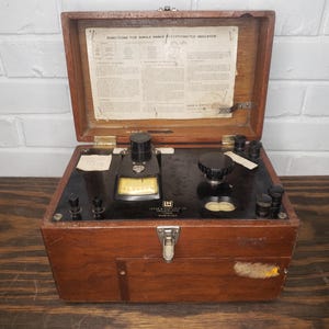 May include: A vintage wooden box containing a single-range potentiometer indicator. The box is brown with a brass latch. The indicator has a black face with a white dial and a black knob. The box has a label that reads "Instructions for Single Range Potentiometer Indicator".