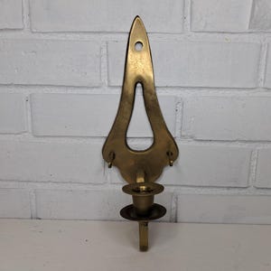 May include: A vintage brass wall sconce with a decorative, elongated design. The sconce features a candle holder and a mounting bracket. The brass has a warm, aged patina, and the sconce is mounted on a white brick wall.
