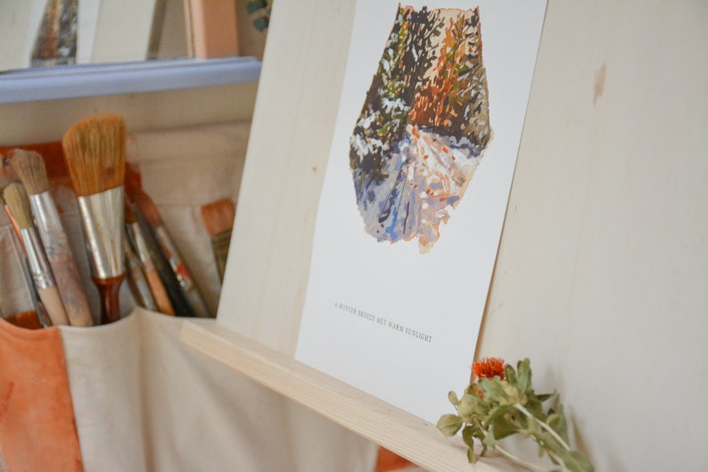 May include: A white framed print of a watercolor painting of a snowy forest path with the text "A winter breeze met with warm sunlight".