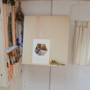 May include: A white wooden easel with a framed print of a forest path. The easel is mounted on a wall with a shelf underneath. A dried flower arrangement is on the shelf.
