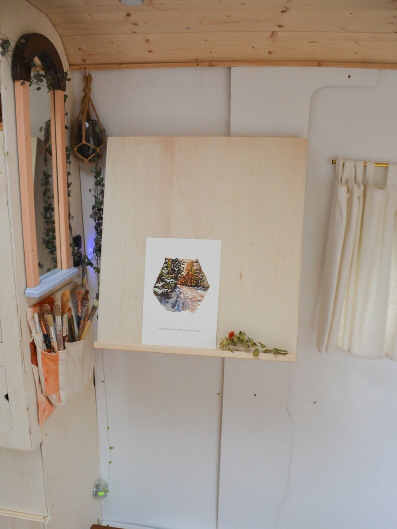 May include: A white wooden easel with a framed print of a forest path. The easel is mounted on a wall with a shelf underneath. A dried flower arrangement is on the shelf.