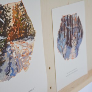 May include: Two watercolor paintings of winter scenes. The first painting shows a snowy path through a forest with trees on either side. The second painting shows a similar scene, but with a more blue color palette. Both paintings have a rough, textured edge.